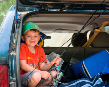 Un jeune enfant prépare sa canne à pêche bien installé dans le coffre arrière ouvert de la voiture.