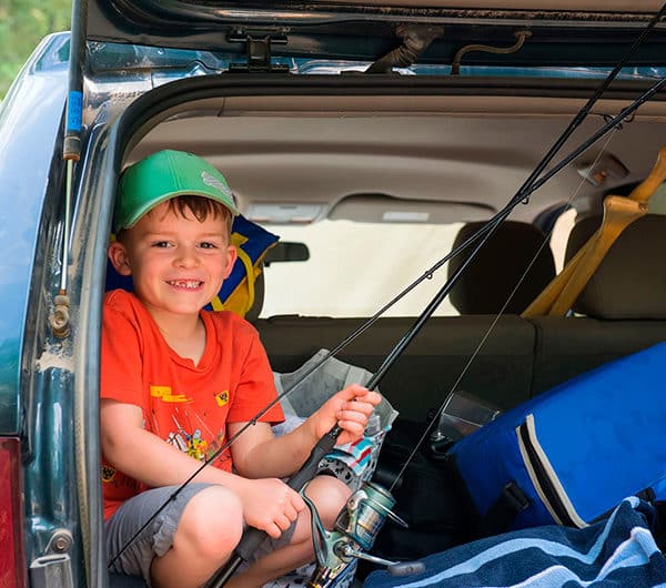 Un jeune enfant prépare sa canne à pêche bien installé dans le coffre arrière ouvert de la voiture.