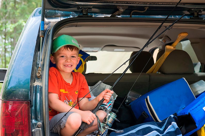 Un jeune enfant prépare sa canne à pêche bien installé dans le coffre arrière ouvert de la voiture.